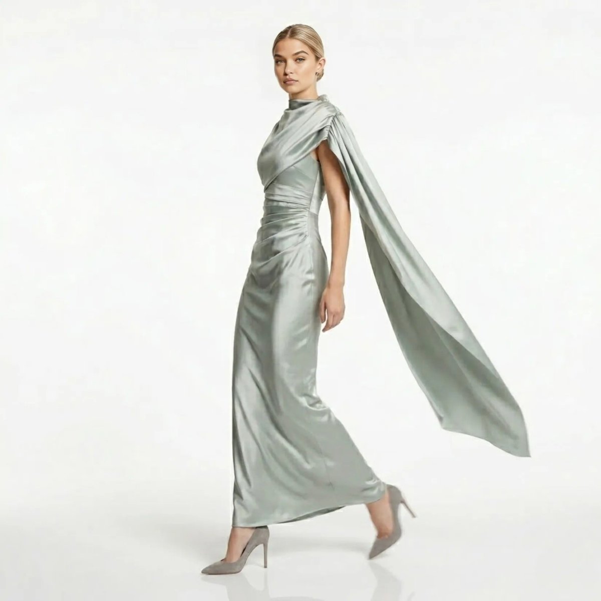 Side profile of model walking in the Kouvr sage green satin formal evening gown with flowing single-shoulder cape detail.