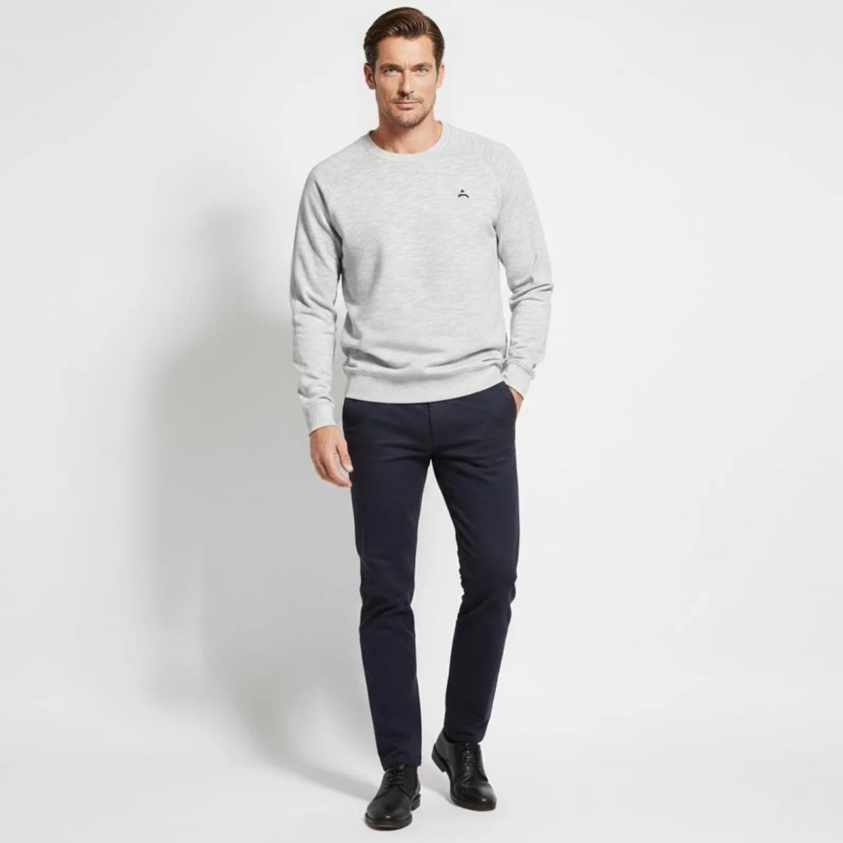 Male model wearing the Kouvr Deacon Crewneck Sweatshirt in heather light grey, styled with dark navy trousers and black shoes, against a white background.