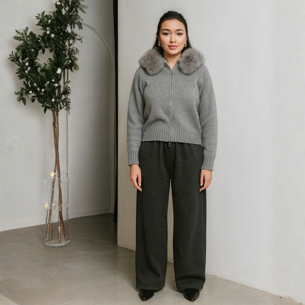 Woman wearing a gray kouvr sweater with fur collar and dark pants standing indoors.