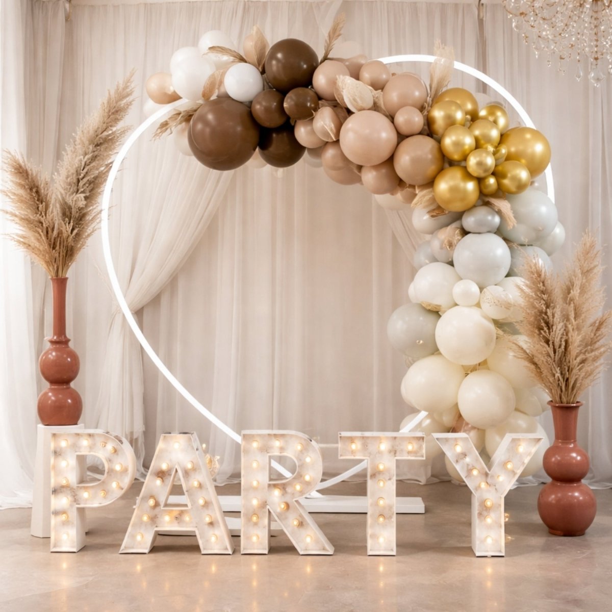 Large heart-shaped party backdrop frame styled with a neutral tone balloon garland, pampas grass, and light-up party letters.