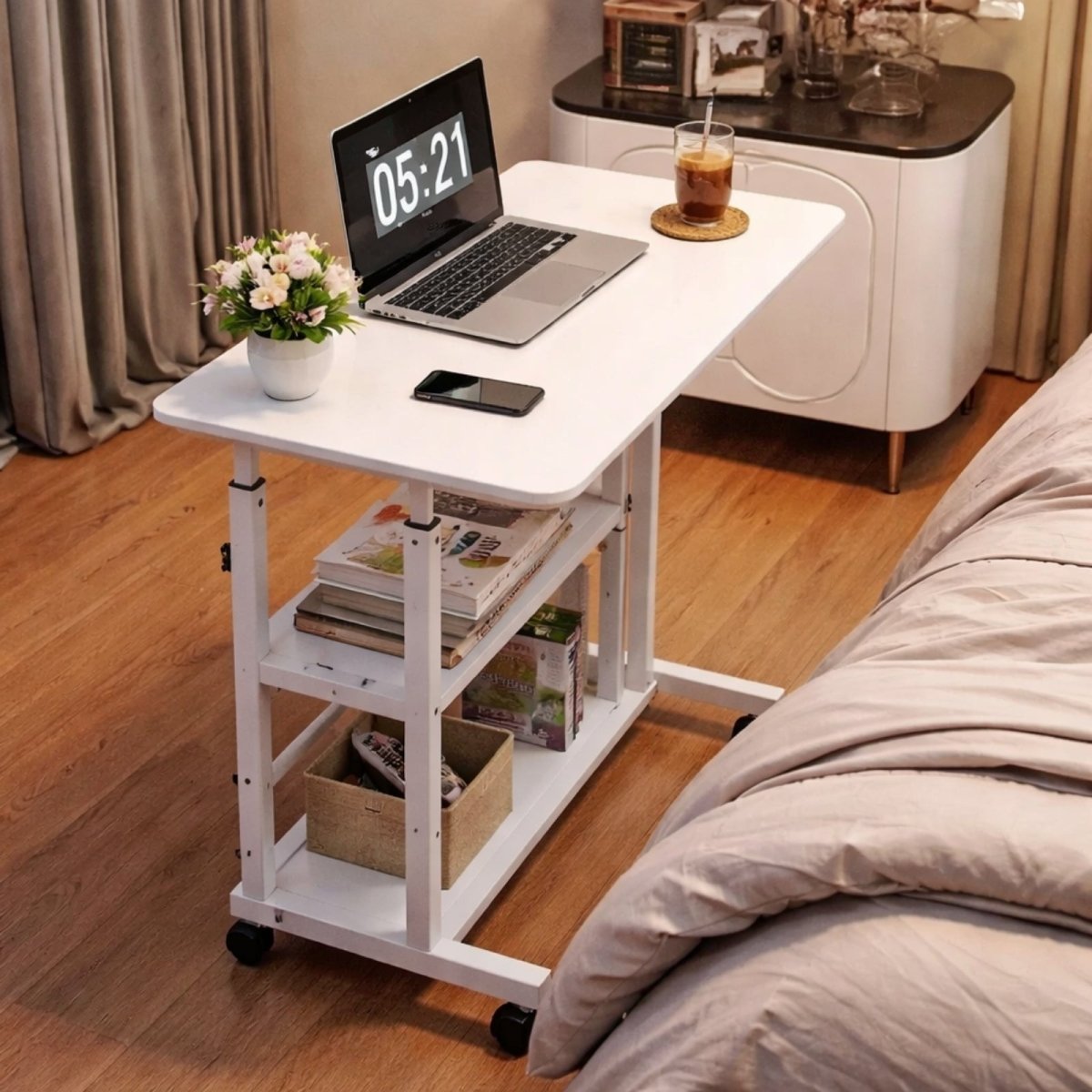 The Kouvr height-adjustable table used as a modern bedside table next to a bed, holding a laptop, drink, and a small vase of flowers.