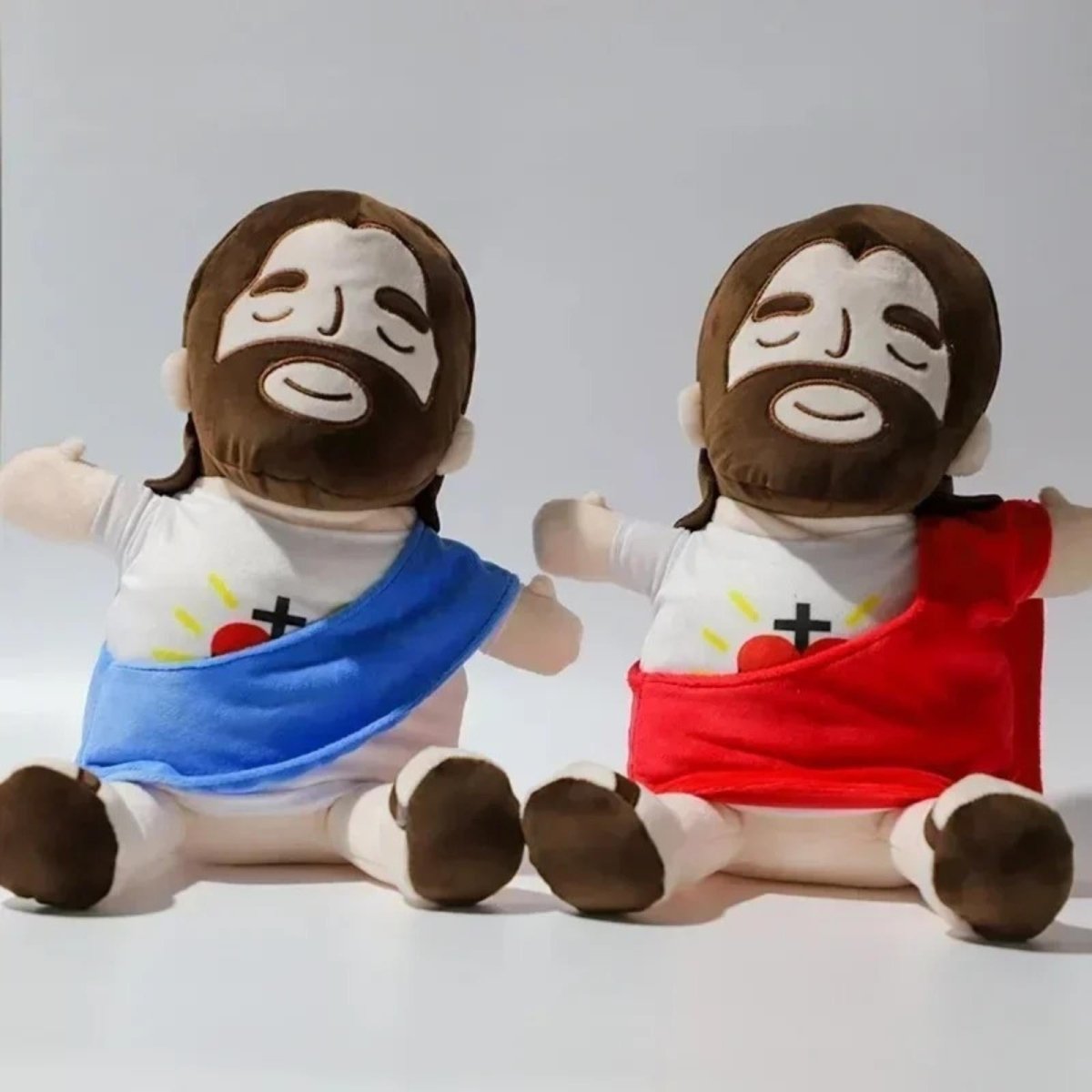 Two Kouvr Jesus plush dolls sitting side-by-side, one featuring a blue sash and the other a red sash representing the Divine Mercy.