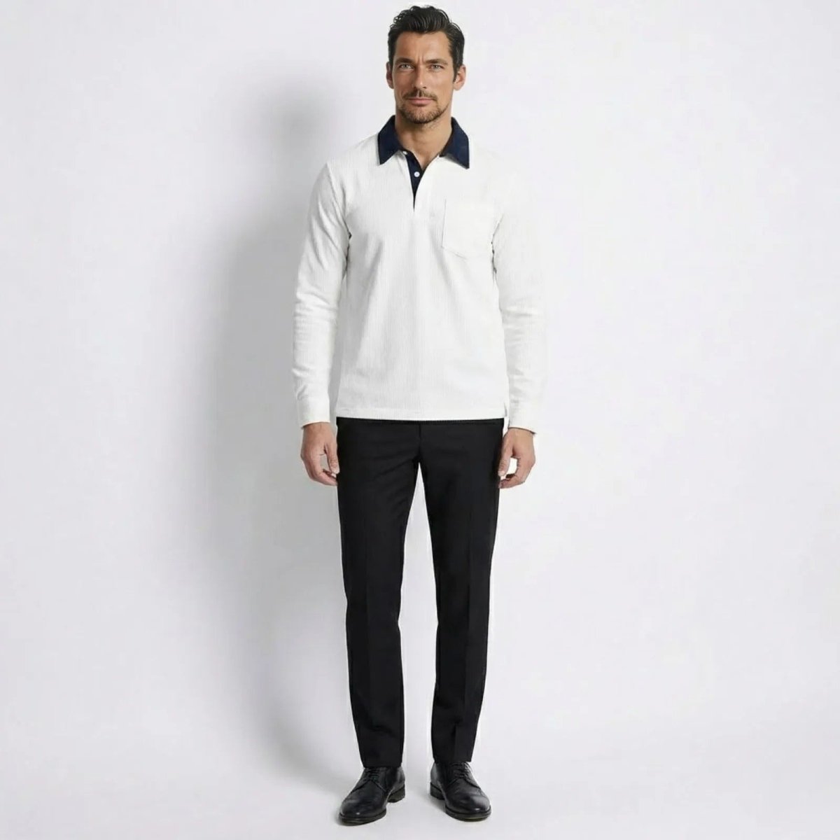 Model facing forward wearing the Kouvr men's white corduroy long sleeve polo shirt with a navy contrast collar, styled with black trousers.