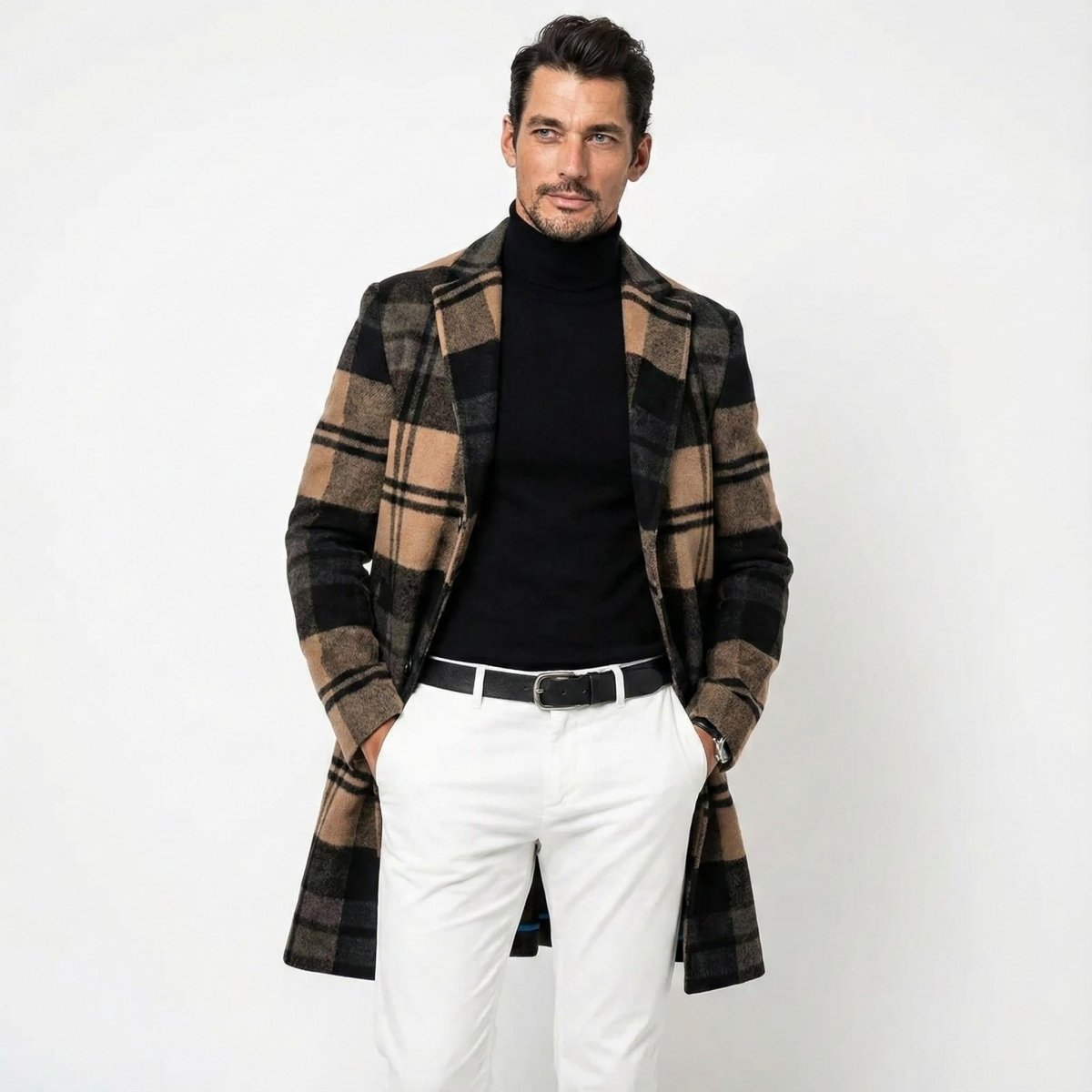 A male model wears the Kouvr tan and black plaid wool-blend overcoat, styled with a black turtleneck and white pants against a white background.