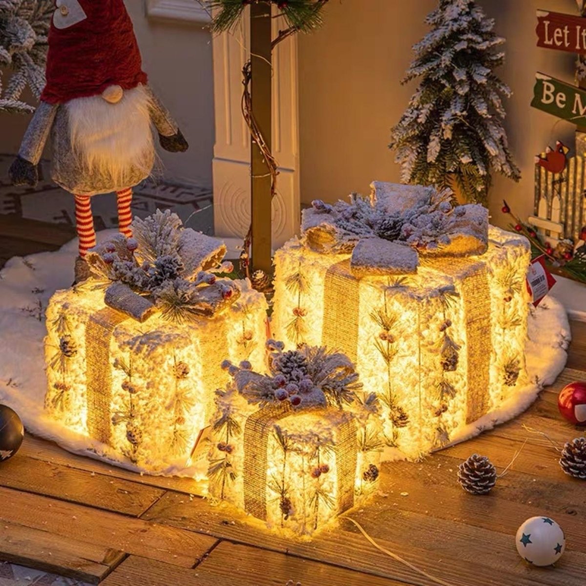 A trio of lighted burlap gift boxes used as outdoor holiday decor on a wooden porch next to a Christmas gnome and a small frosted tree.