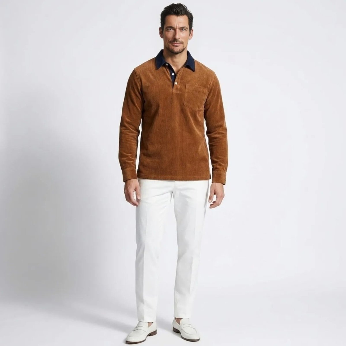 Model wearing the Kouvr Zain long sleeve corduroy polo shirt in Orange with white trousers, showing the contrast navy collar.