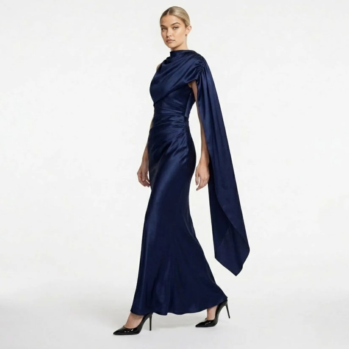 Side profile of a female model wearing the navy blue ruched satin formal dress by Kouvr showcasing the cape detail.