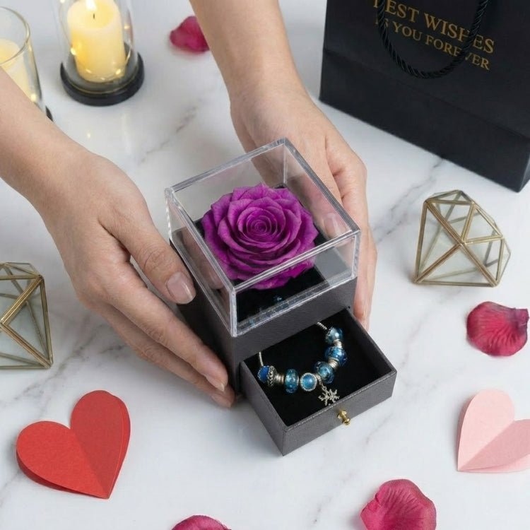 Hands holding a purple preserved rose gift box with a clear acrylic lid and an open drawer revealing a blue bead charm bracelet.