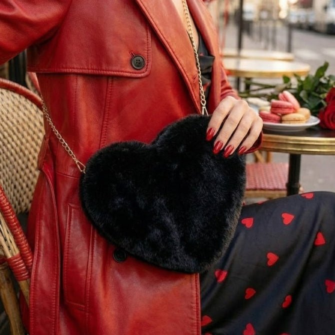 Close-up of a woman wearing a red leather trench coat and a black heart-print dress, carrying a black furry heart-shaped crossbody bag at a Parisian cafe.