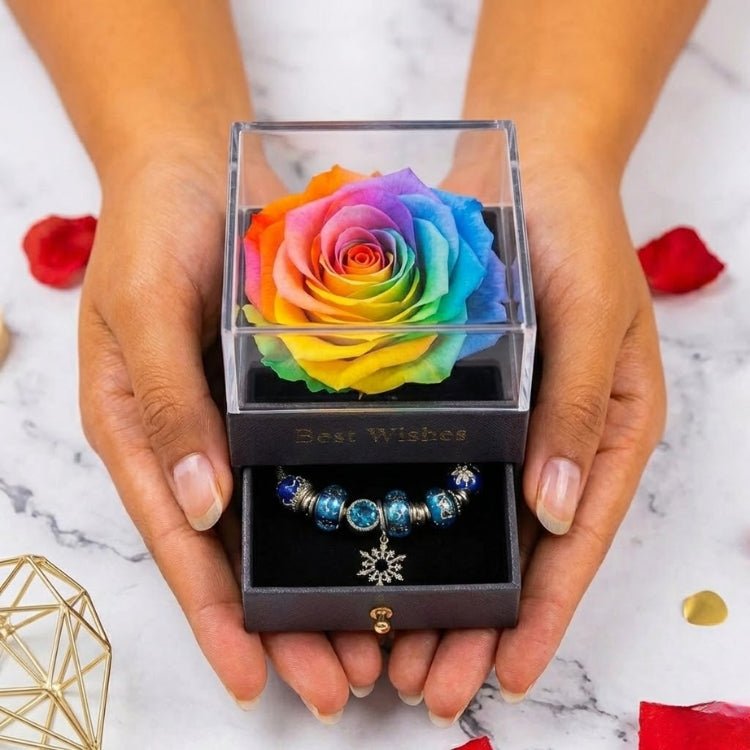 Hands holding a multicolor rainbow eternal rose display box with a clear acrylic lid and a hidden drawer containing a blue bead bracelet.