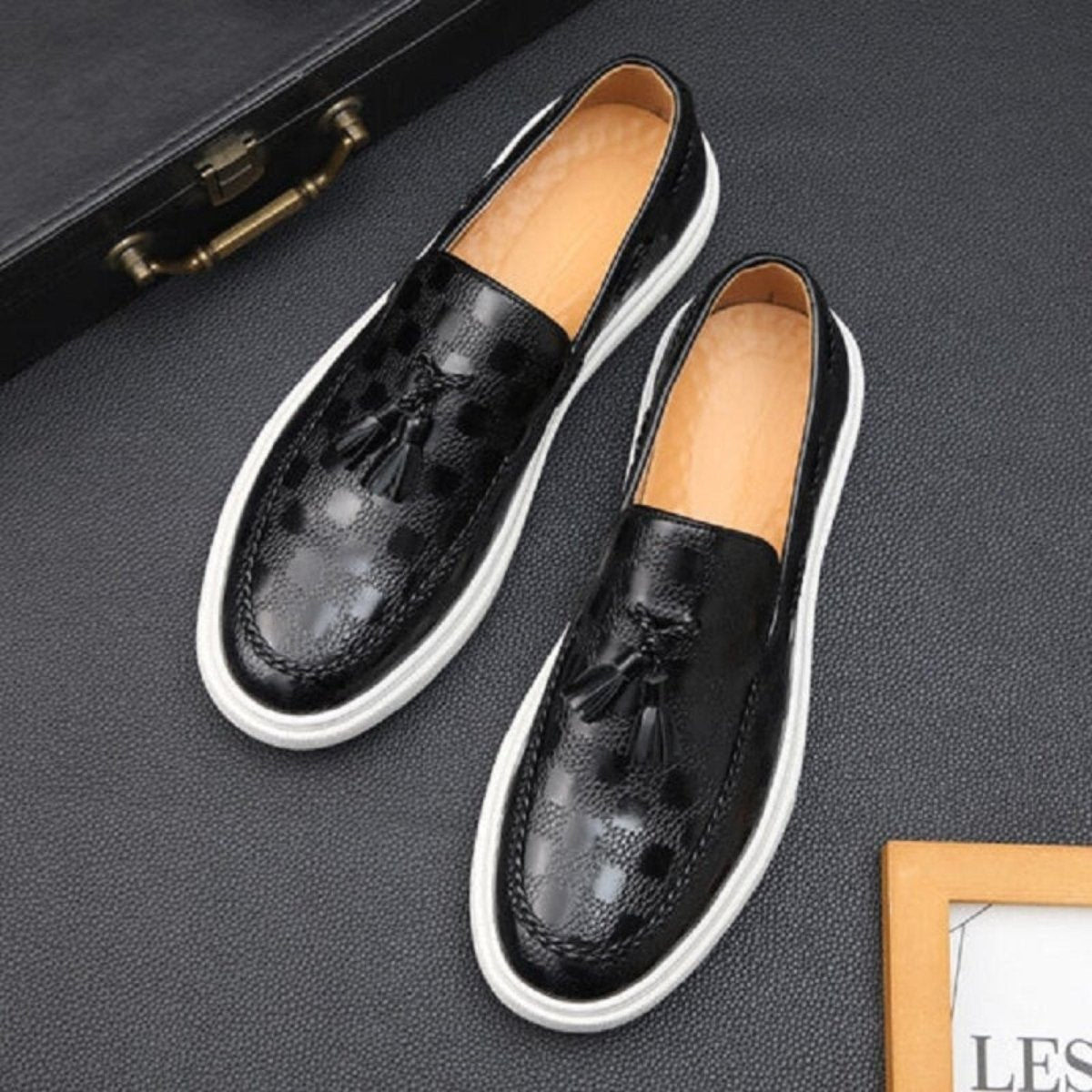 A pair of Kouvr Carlo men's black embossed leather tassel loafers with a distinctive checker pattern and white sole, shown from a top-down angle. 