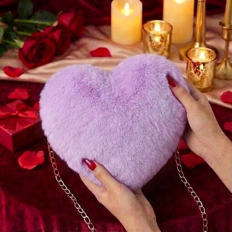 Close-up of a woman's hands with red nails holding a soft lavender faux fur heart-shaped clutch bag in a romantic setting with candles and roses.

