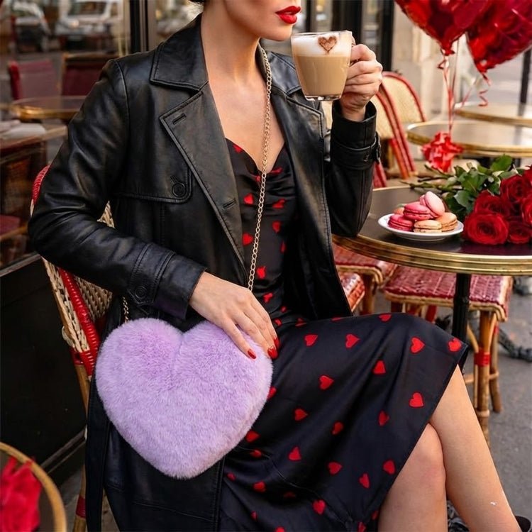 Woman in a heart-print dress and black jacket holding a cup of coffee at an outdoor cafe, with a lavender faux fur heart-shaped purse on her lap.