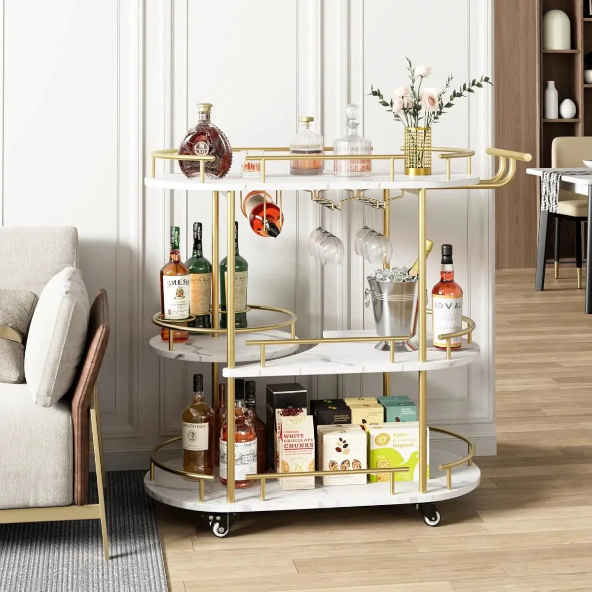 Kouvr luxury gold rolling bar cart with three white marble shelves, filled with liquor bottles and glassware, featuring a handle and wine rack.