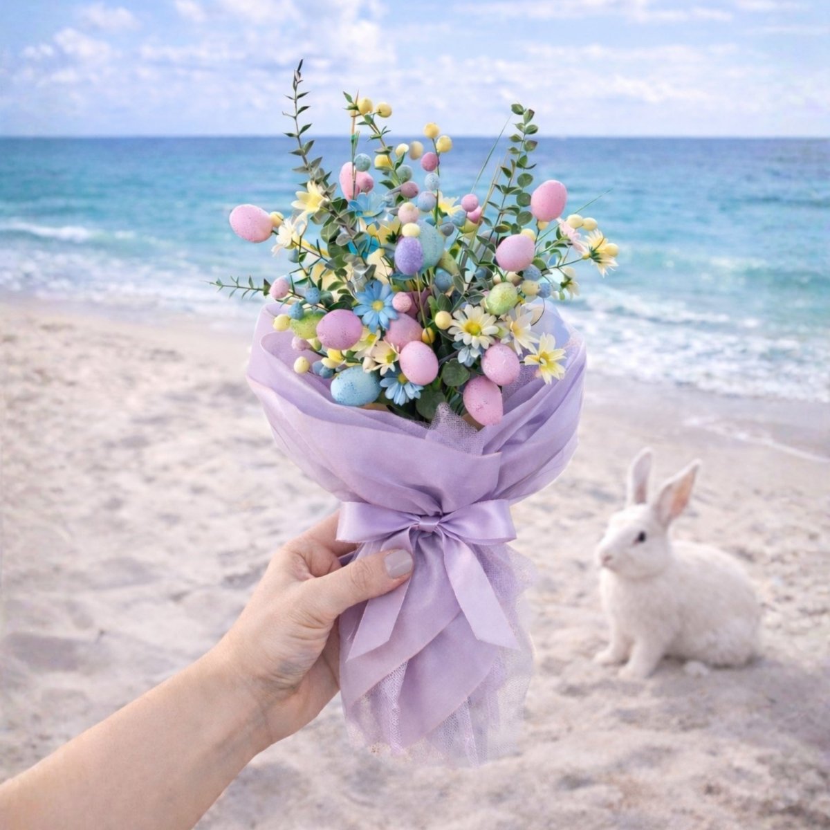 A hand holding a purple-wrapped Easter egg bouquet on a sandy beach with a white rabbit and the ocean behind it.