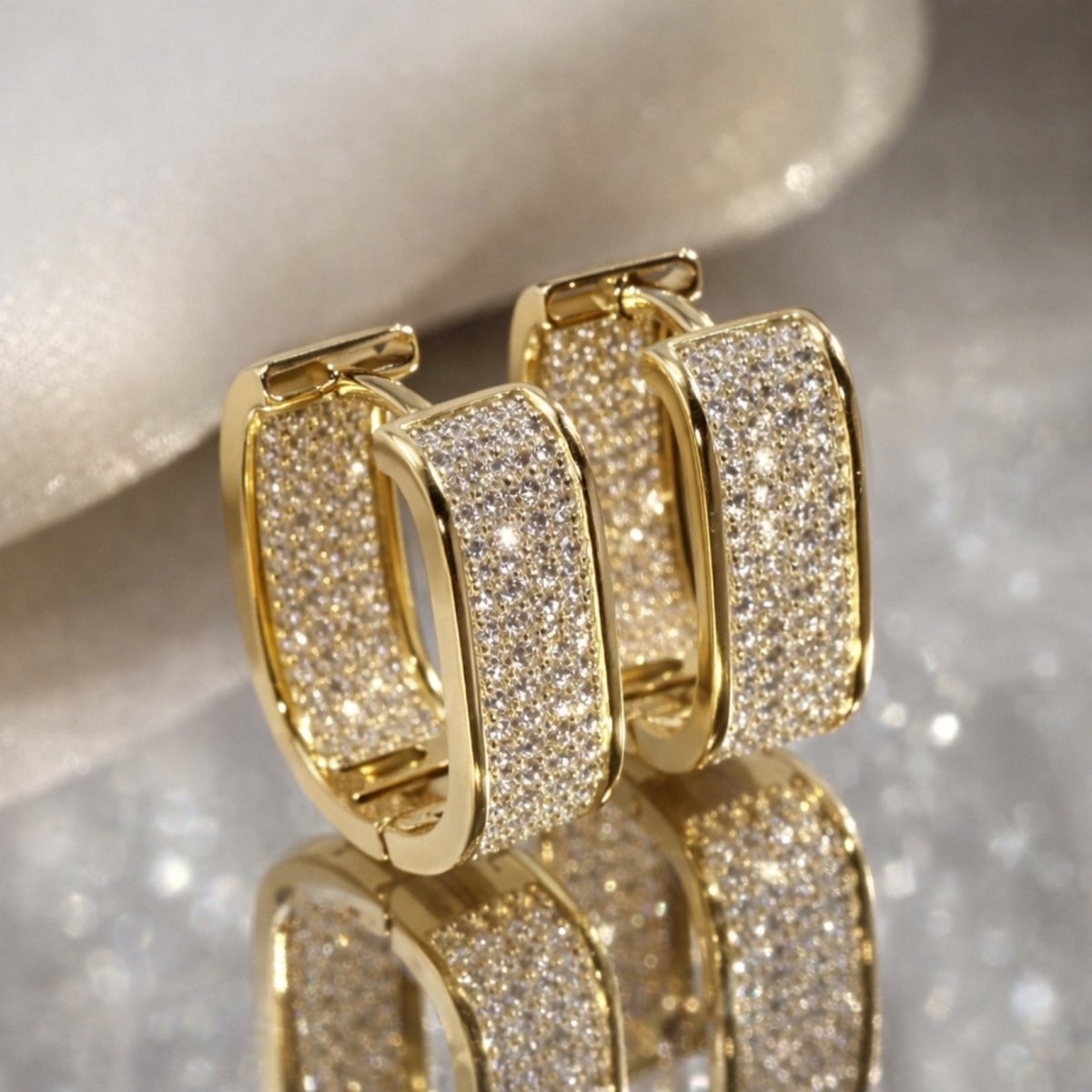 A pair of gold Kouvr Salome rectangular pavé hoop earrings with white crystals, styled on a reflective, sparkling surface with a silk background.