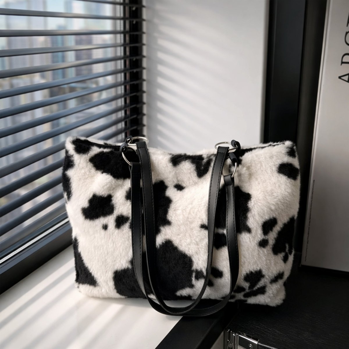 The Kouvr Sorooya cow print tote bag sitting on a white windowsill, highlighting its fluffy texture and silver hardware details. 