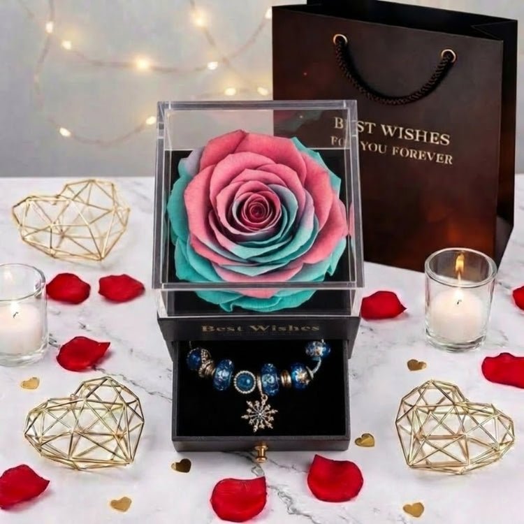 Top-down view of a vibrant pink and teal gradient preserved rose in a black gift box, displayed with a snowflake charm bracelet and romantic decor.
