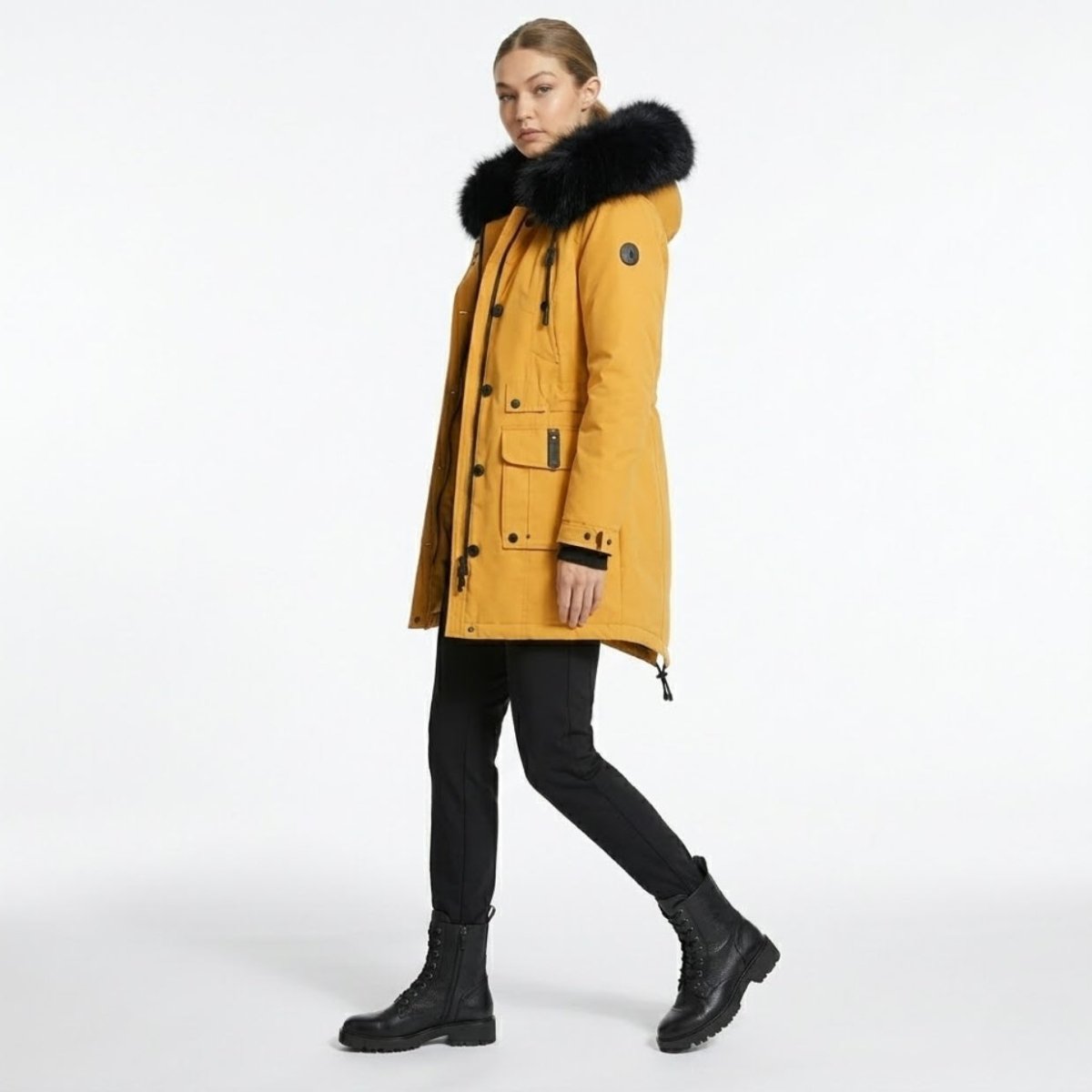 Side profile walking shot of a woman modeling a Kouvr mustard yellow insulated winter parka jacket showing the tailored mid-length fit and oversized black fur hood.