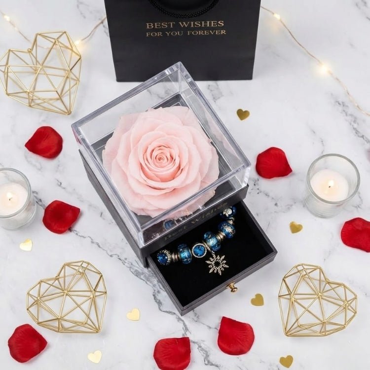 Top-down view of a pale pink forever rose in a luxury gift box surrounded by red rose petals, candles, and gold geometric hearts.