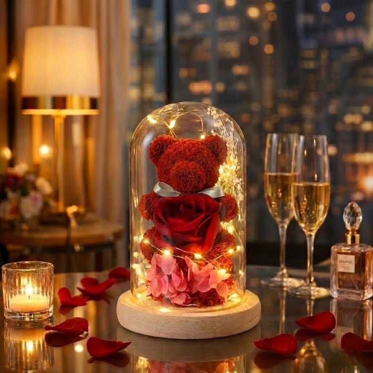 Classic red eternal rose teddy bear in a glass cloche with warm fairy lights and scattered red rose petals.