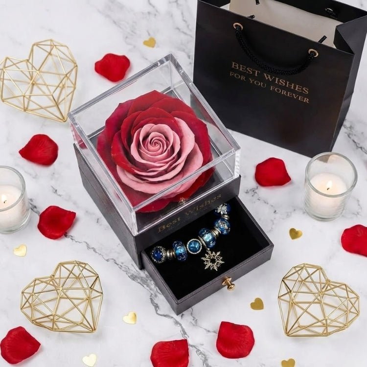 A unique two-tone red and pink eternal rose in a black jewelry box with a snowflake charm bracelet, displayed next to a black "Best Wishes" gift bag.